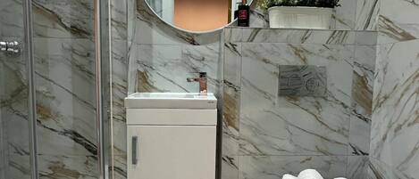 A bathroom with a glass shower enclosure, a white sink, a round mirror, a toilet with towels, and a potted plant on a shelf.