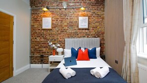 A hotel room with a brick wall, a bed with blue and red pillows, a laptop on the bed, and a window with curtains. - Farringdon Rooms (London)