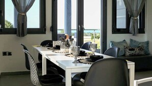 Un coin repas avec une table blanche dressée pour quatre personnes, un canapé avec des coussins et de grandes fenêtres dotées de rideaux.