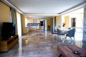 Uma sala de estar ampla com TV de tela plana, um móvel para entretenimento de madeira, uma mesa de jantar com cadeiras e uma cozinha ao fundo.