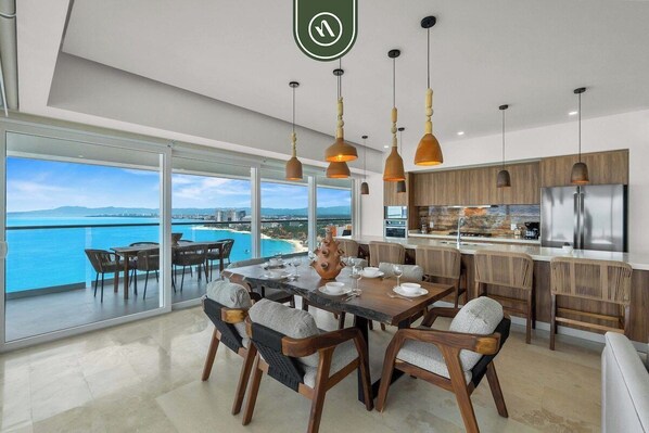 A modern kitchen with a view of the ocean, large windows, and a dining area with a table and chairs.