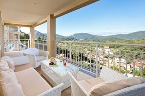 A balcony with white wicker furniture, a glass-top table with fruits and drinks, and a scenic view of green hills and buildings. - Pantokrator Private Pool Gem - Lux Villa in Corfu (Agios Mattheos)