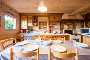 A wooden cabin interior with a dining table set for four, wooden cabinets, a kitchen area with appliances, and a large window.