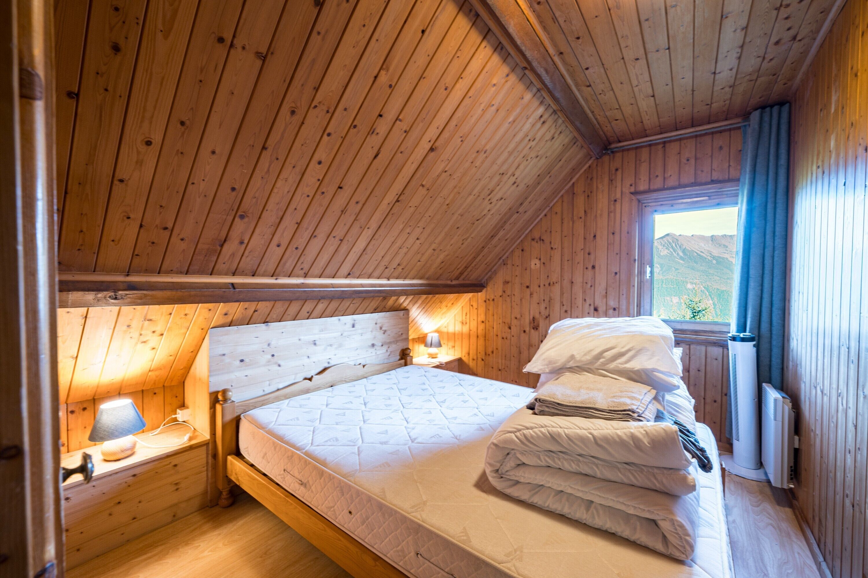 Ein Schlafzimmer einer Holzhütte mit einem Einzelbett, einem Nachttisch mit Lampe und einem Fenster mit Blick auf die Berge.