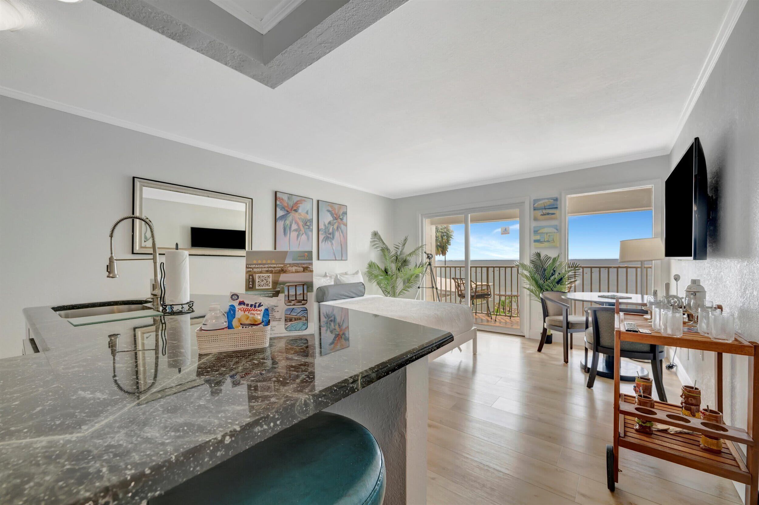 A modern kitchen with a marble countertop, a sink, and a large mirror. There are framed pictures on the wall, a dining area with a table and chairs, and a balcony with a view of the ocean.