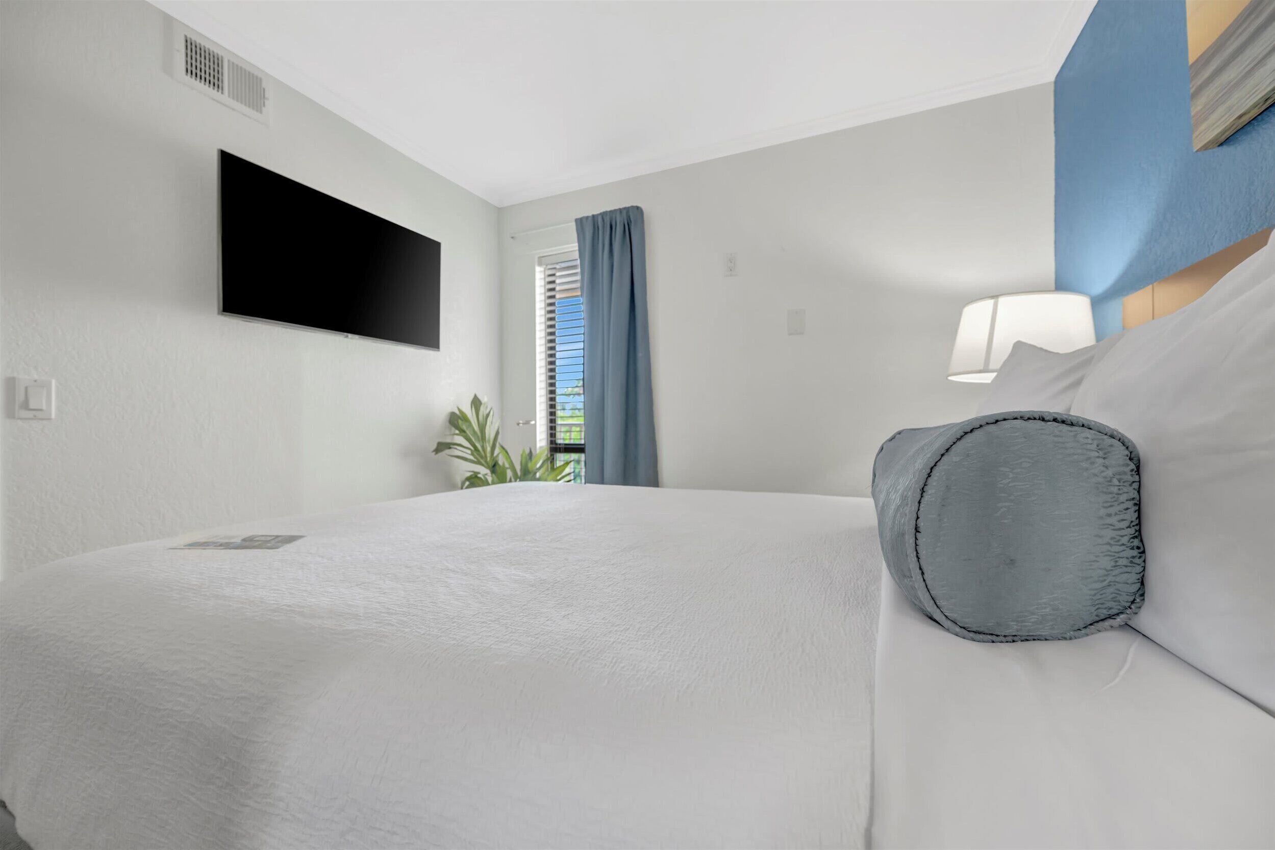 Una habitación de hotel con una cama, un televisor de pantalla plana montado en la pared, una ventana con cortinas azules y una lámpara de noche.