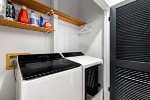 A compact laundry area with a washing machine and dryer, a wooden shelf with cleaning supplies, and a black roller shutter door.