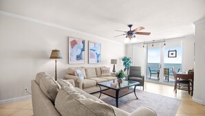 A living room with a sofa, coffee table, and a ceiling fan. There are two paintings on the wall and a view of the ocean through the sliding glass door. - NEW LISTING!!! Seaspray East 922 - Beach Front - HASSLE-FREE CHECK OUT (Perdido Key)
