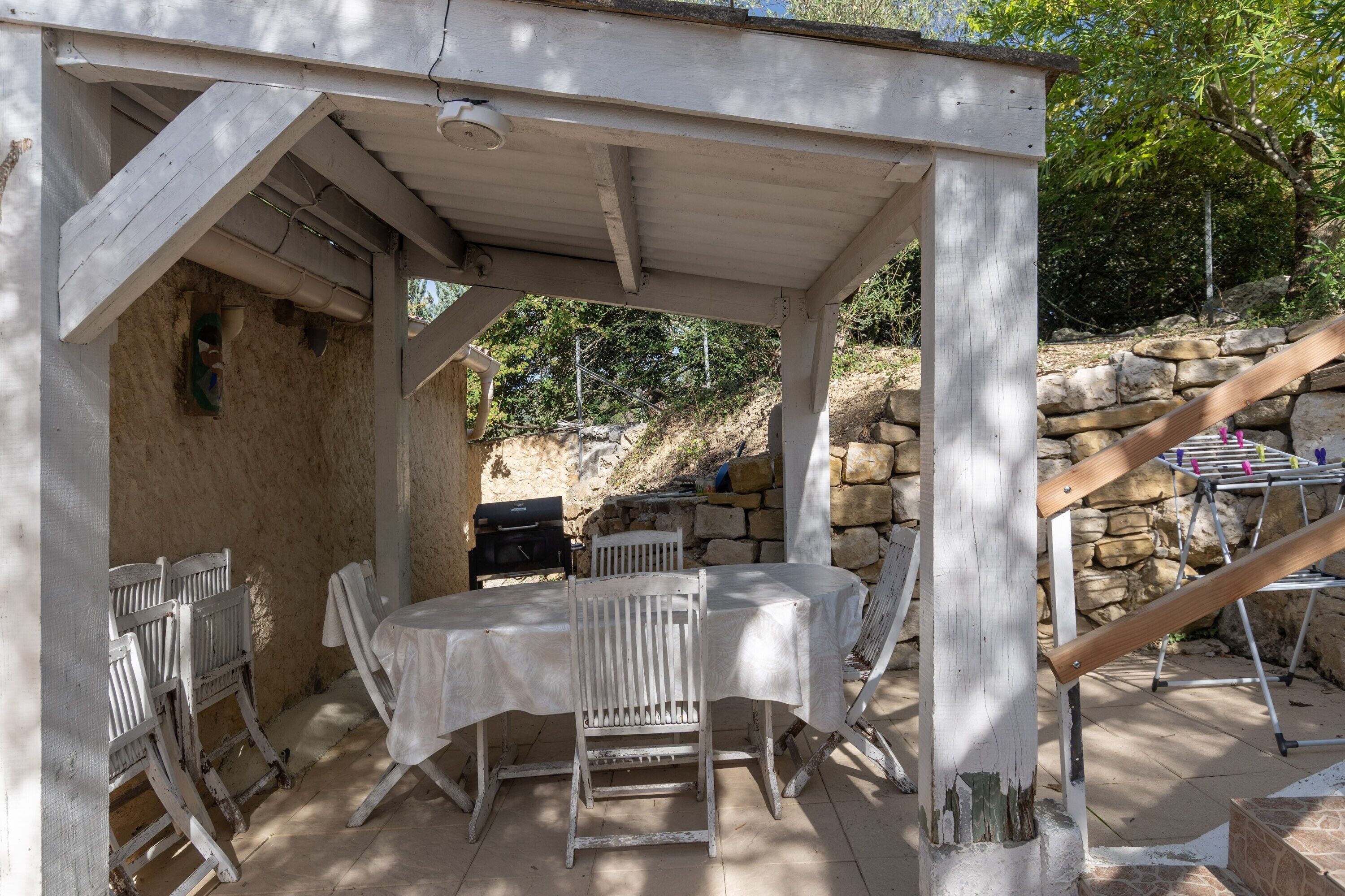Um pátio externo com mesa, cadeiras, churrasqueira e um muro de pedra ao fundo.