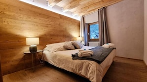 Une chambre avec des murs en bois, un lit recouvert d’une couette beige et grise, une lampe de chevet et une fenêtre avec des rideaux.