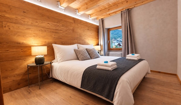 A hotel room with a wooden ceiling, a bed with white linens, a nightstand with a lamp, and a window with a view of a village. - Alpine Chic Retreat (Courmayeur)