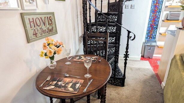Un coin repas avec une table ronde en bois, dressée pour quatre personnes, un vase de fleurs et un escalier orné de rampes noires.