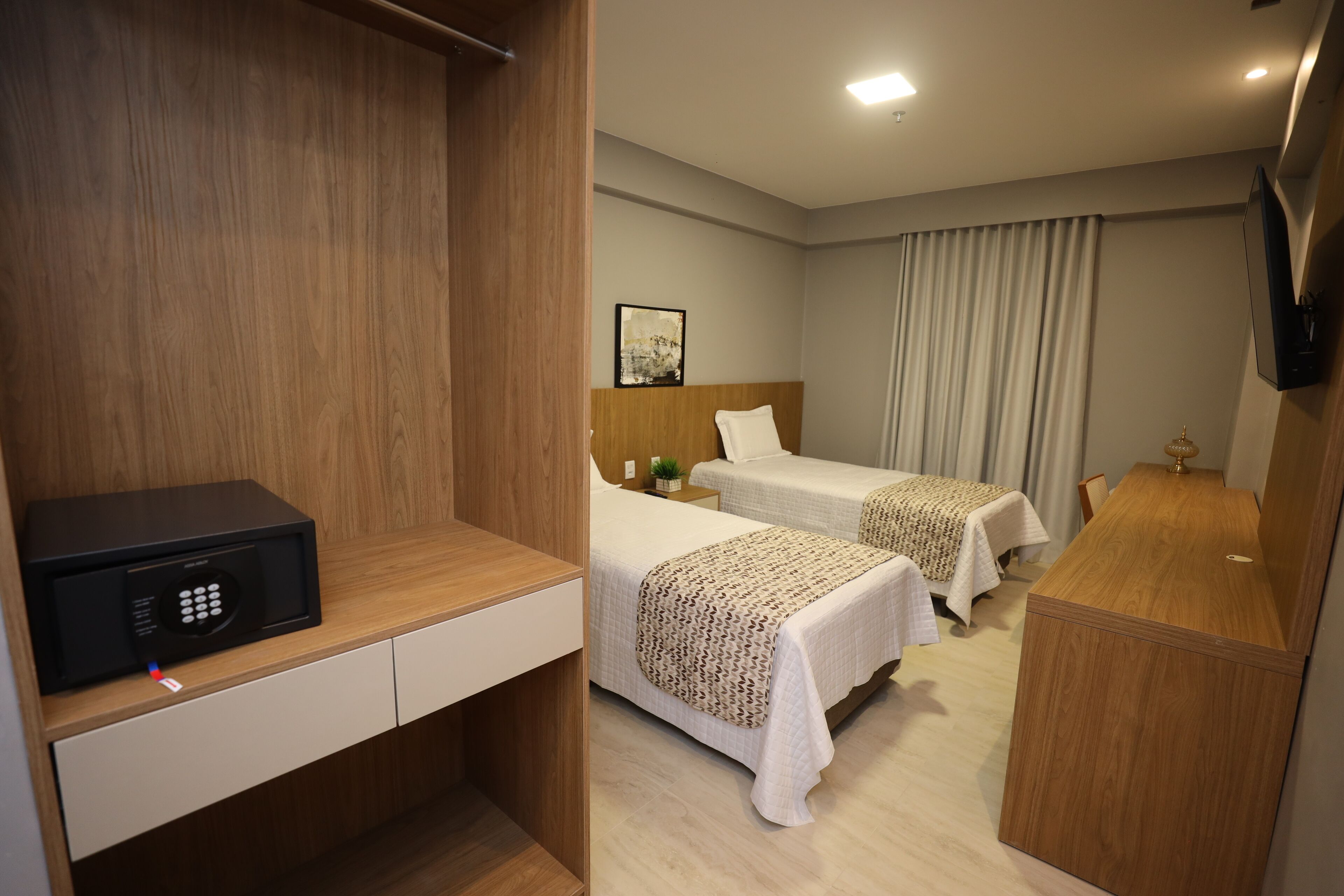 A hotel room with a wooden desk, a microwave, two beds with patterned bedding, a bedside table with a lamp, and a TV mounted on the wall.