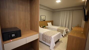 A hotel room with a wooden desk, a microwave, two beds with patterned bedding, a bedside table with a lamp, and a TV mounted on the wall.