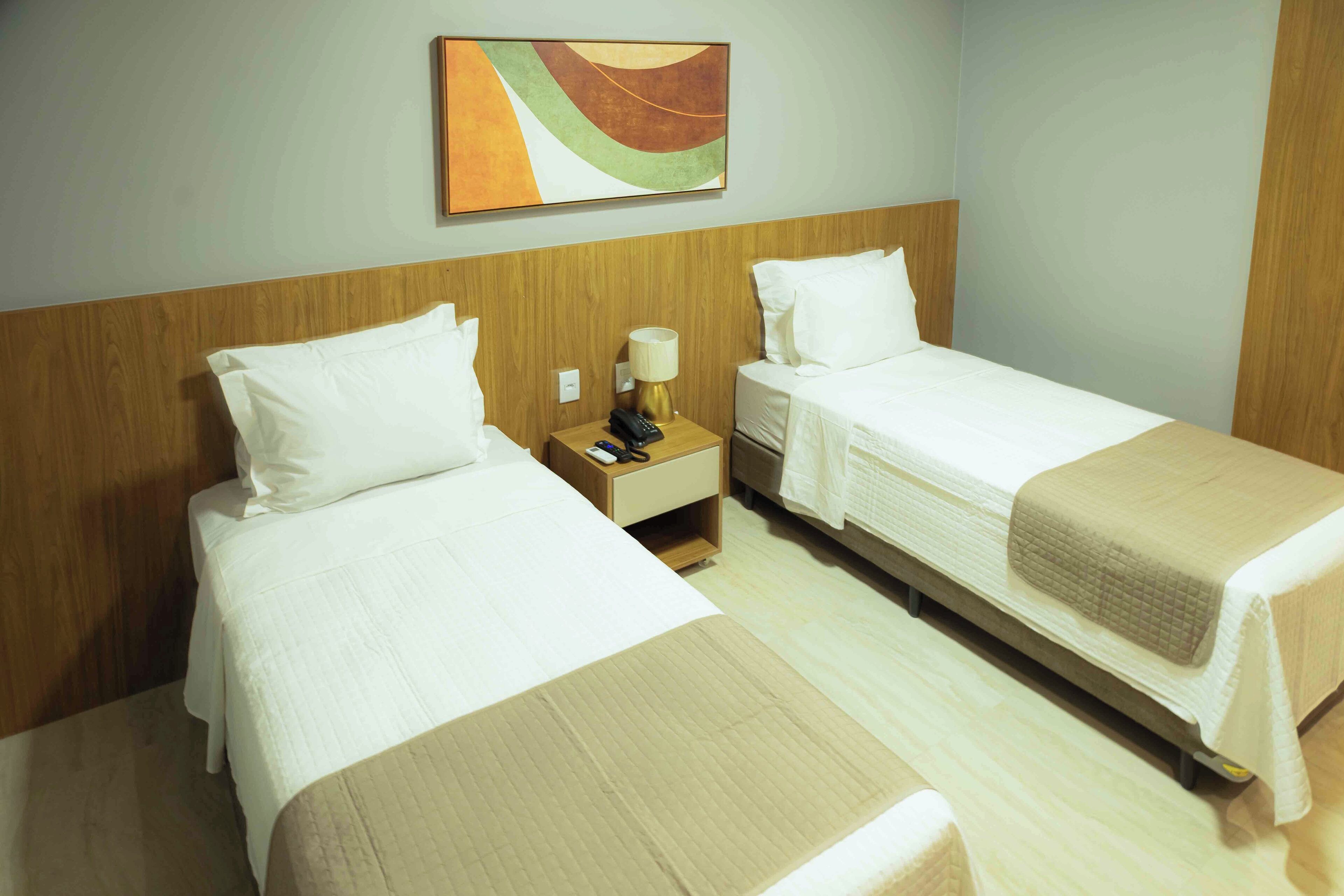 Una habitación de hotel con dos camas individuales, un cabecero de madera, una mesita de noche con lámpara y un cuadro en la pared.