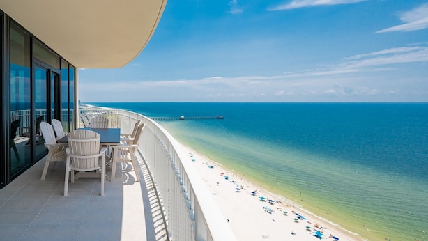 A balcony with a table and chairs overlooking a beach with a pier in the distance. - Phoenix Gulf Shores Ii 2205 4 Bedroom Condo (Gulf Shores)
