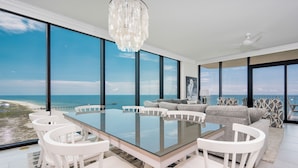 A modern dining area with a glass table and white chairs, overlooking a beach and ocean. - Phoenix Gulf Shores Ii 2205 4 Bedroom Condo (Gulf Shores)