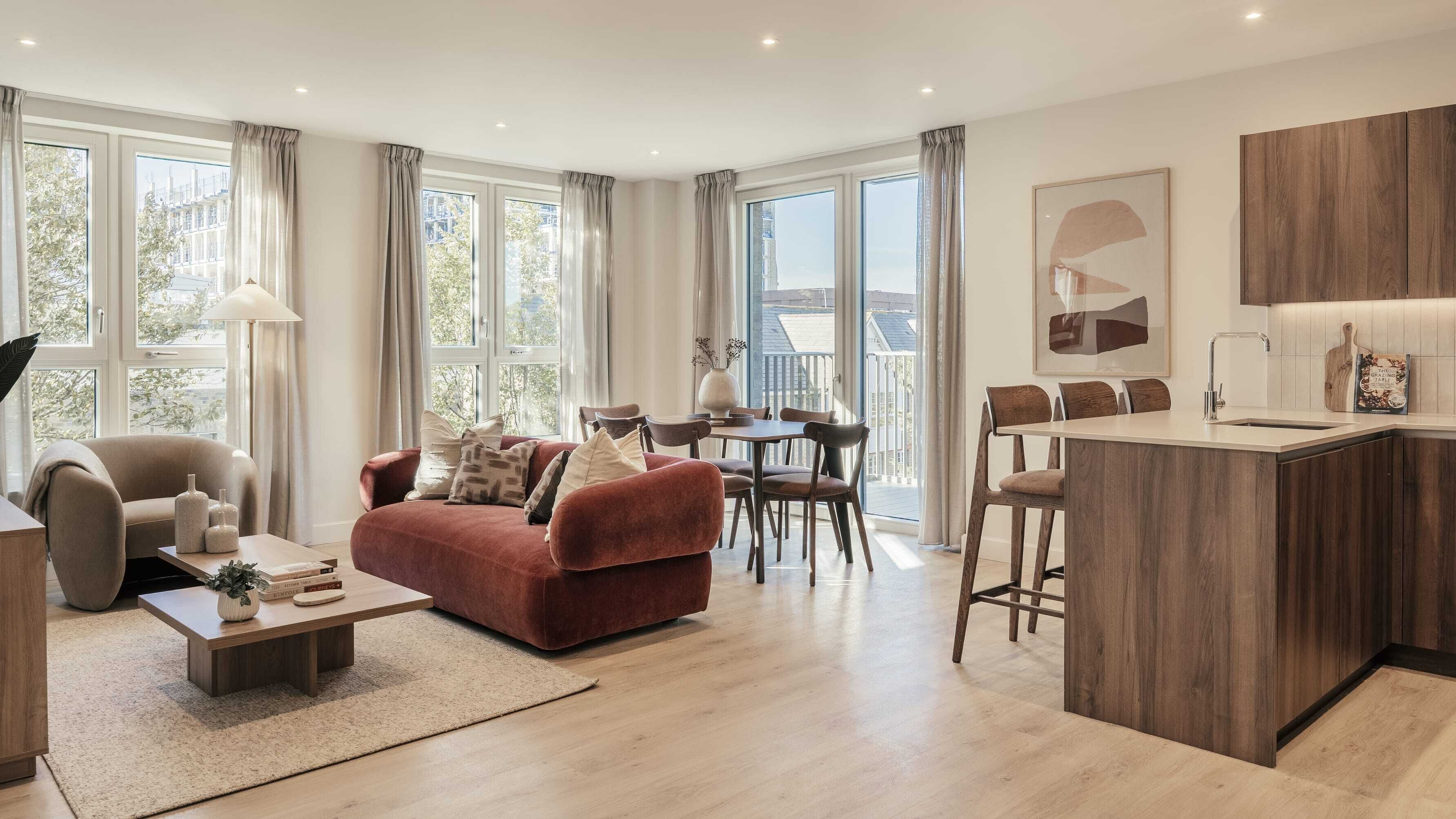 A modern living room with a red sofa, a coffee table, and a dining area with chairs. There is a kitchen with a wooden countertop and a sink. Large windows provide natural light and views of the outside.