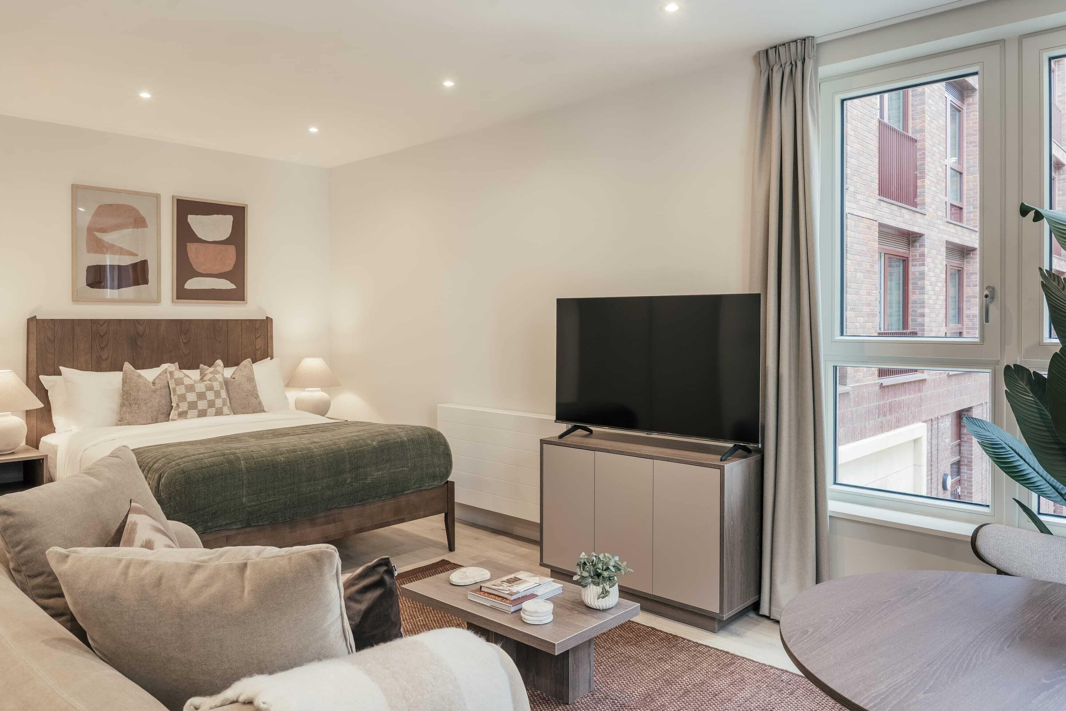 A modern hotel room with a bed, a TV, a sofa, a coffee table, and a window with a view of buildings.