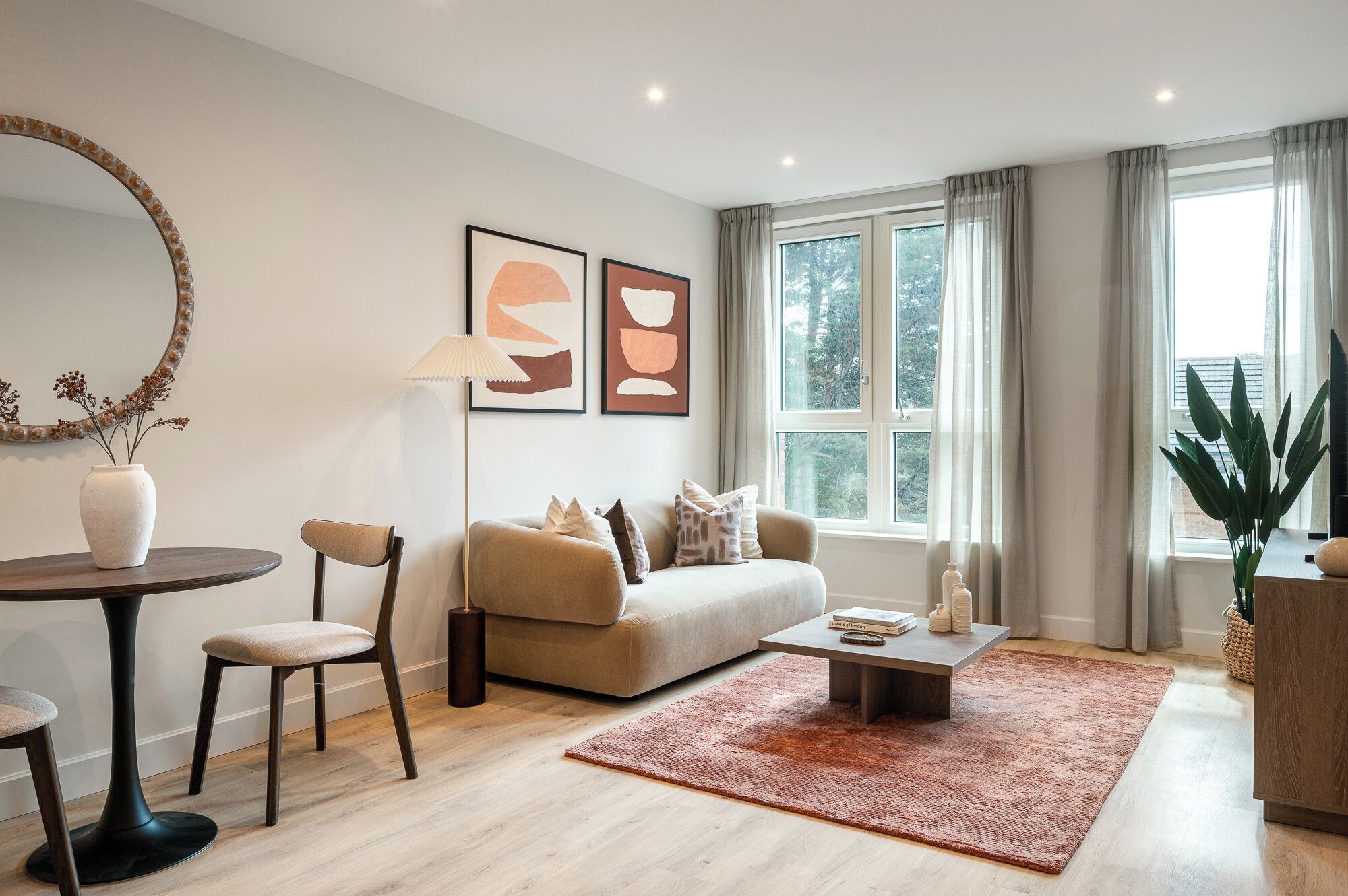 A modern living room with a sofa, coffee table, and chairs. There is a large window with curtains, a round table with a vase, and two framed abstract paintings on the wall.