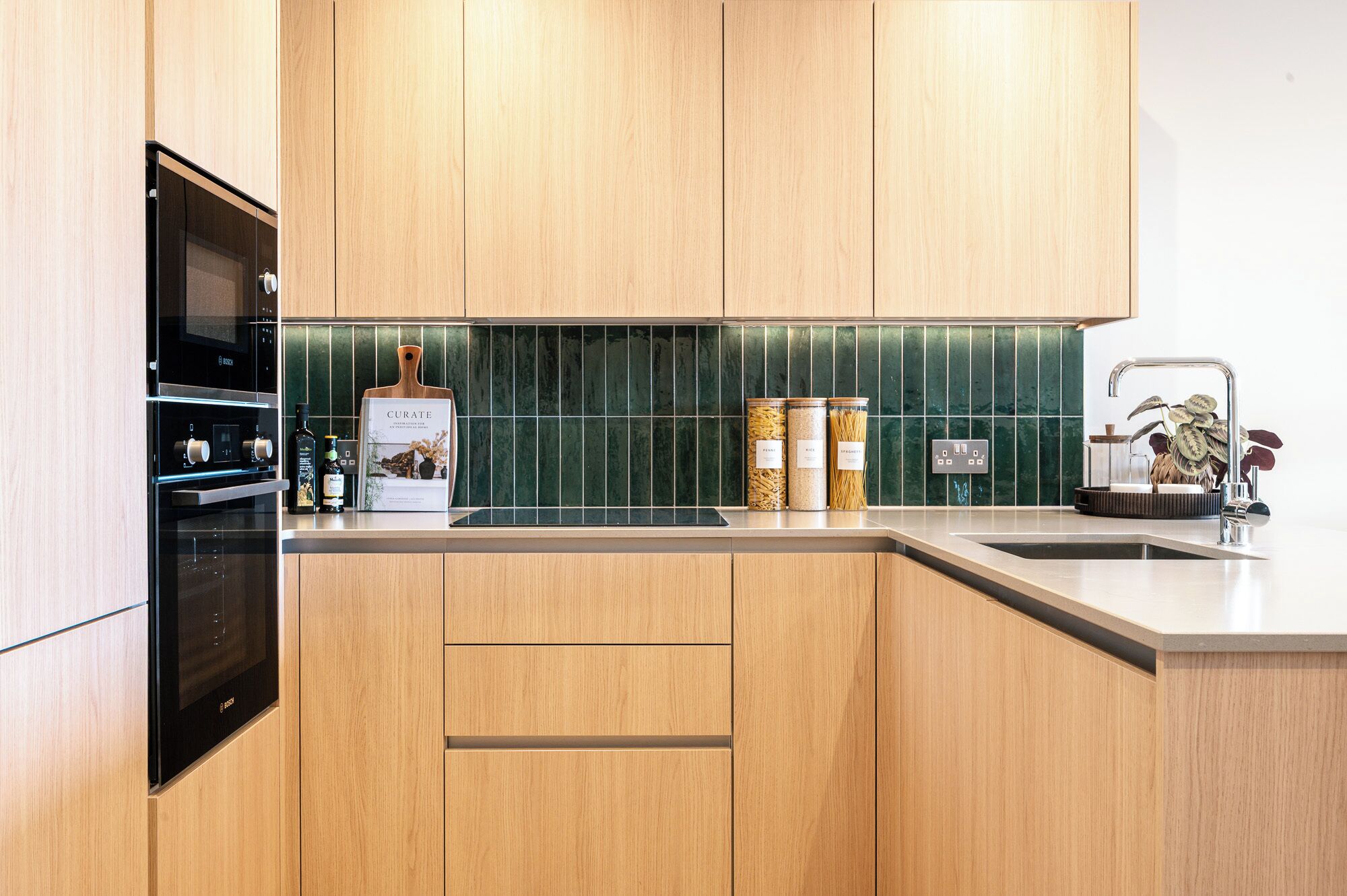 Una cocina moderna con armarios de madera, un horno negro y un salpicadero verde. Hay frascos de pasta y un libro en la encimera.