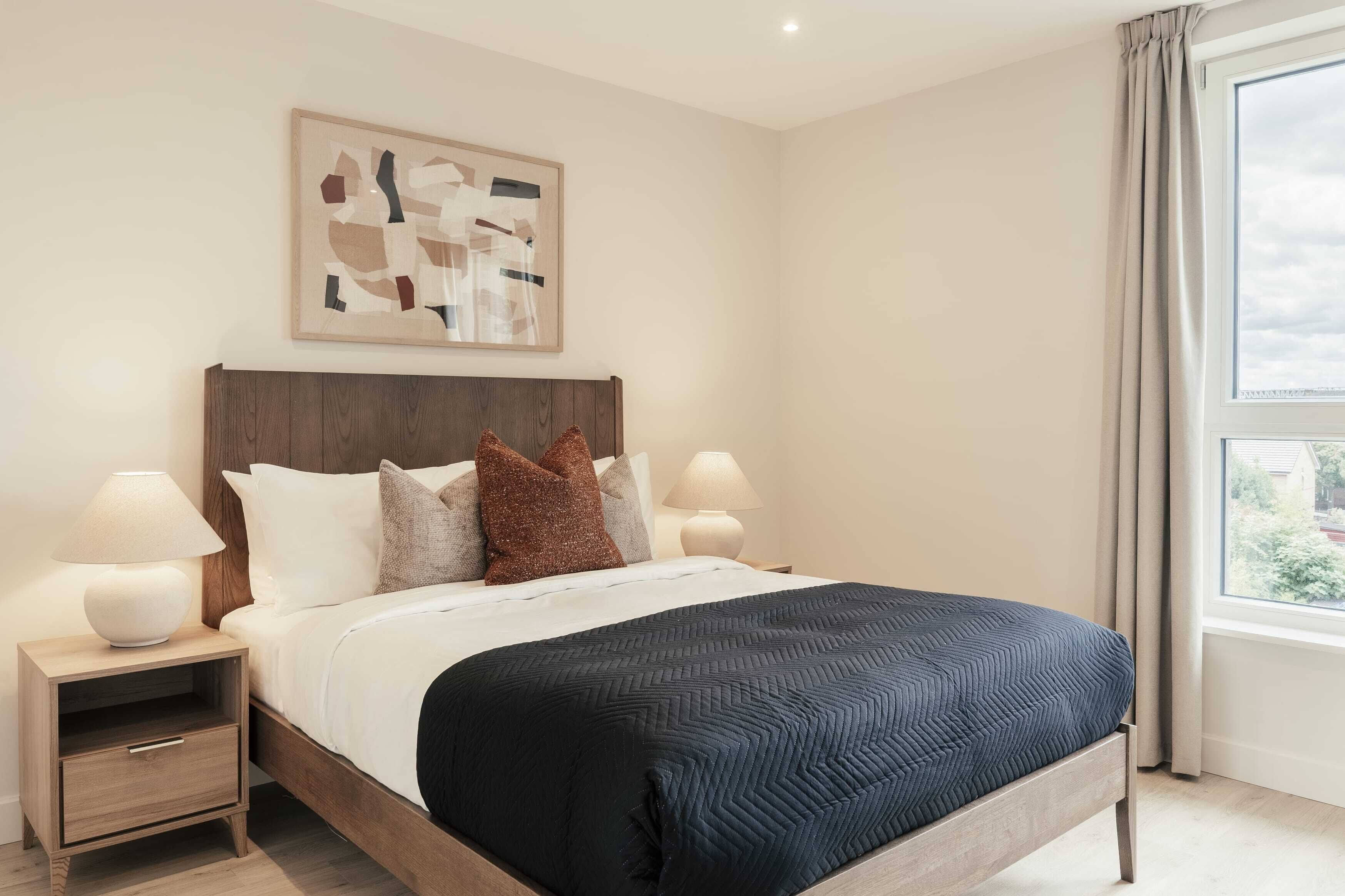 A neatly made bed with a dark quilt, two bedside lamps, a wooden nightstand with a drawer, and a large window with curtains.