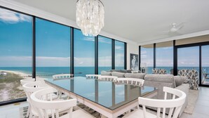A modern dining area with a glass table and white chairs, overlooking a beach and ocean.
