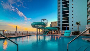 A swimming pool with a water slide, a high-rise building, and a sunset sky.
