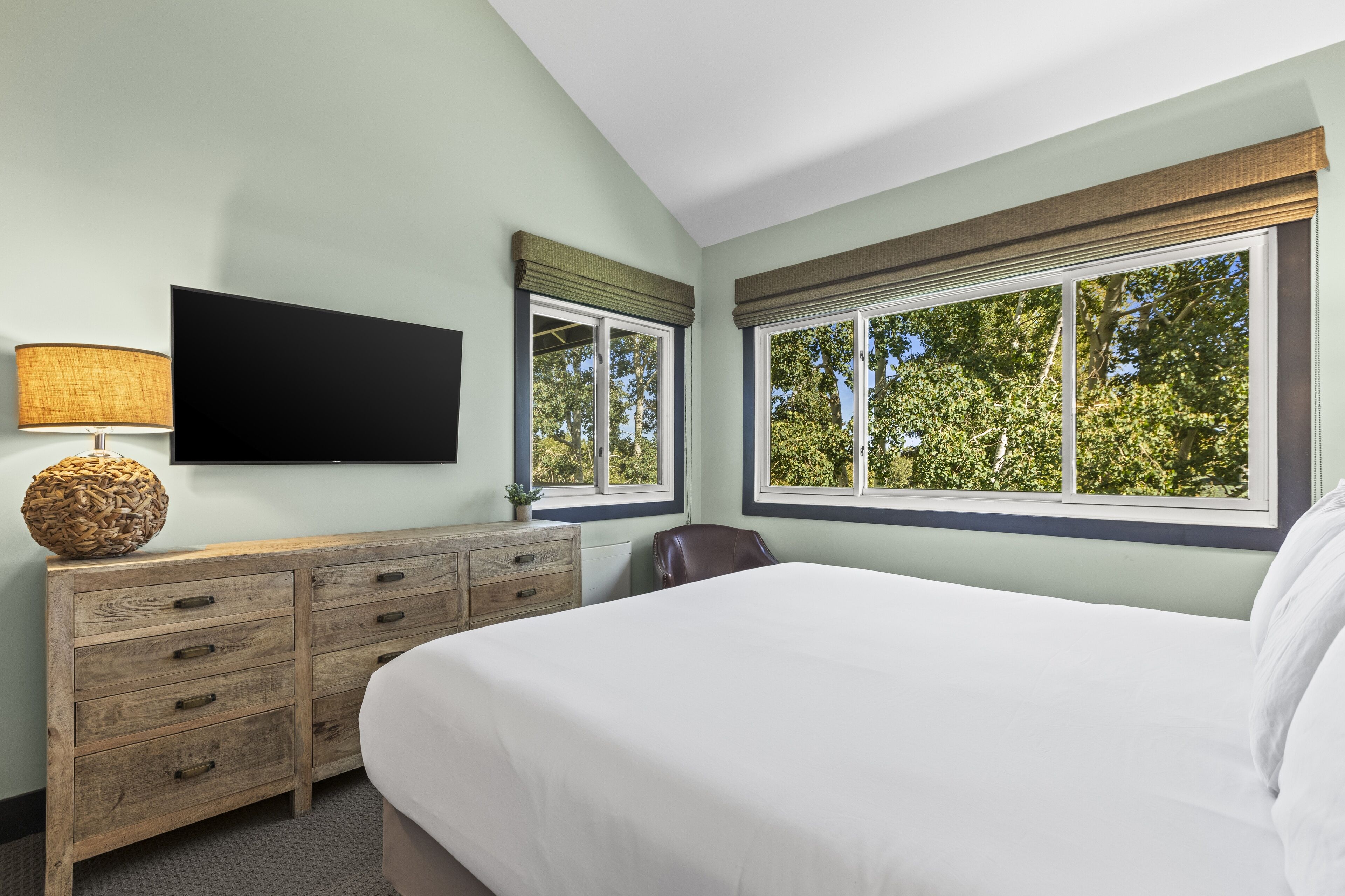 A bedroom with a large bed, a wooden dresser with drawers, a flat-screen TV mounted on the wall, and two windows with blinds.