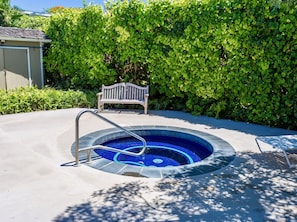 Uma piscina circular com interior azul, rodeada por um deck de concreto, um banco de madeira e uma sebe verde densa.