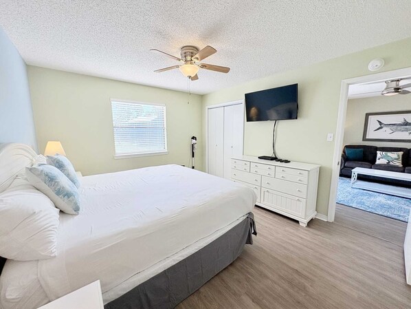 Une chambre à coucher comprenant un lit, un ventilateur de plafond, une télévision, une commode et une porte donnant sur une autre pièce.