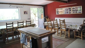 A dining area with wooden chairs and a table, a window with a view of greenery, and a wall with framed pictures.
