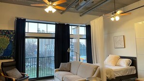 Un salon moderne avec un canapé, un fauteuil et un lit à baldaquin. On y trouve deux ventilateurs de plafond, une grande fenêtre avec des rideaux et un tableau accroché au mur.