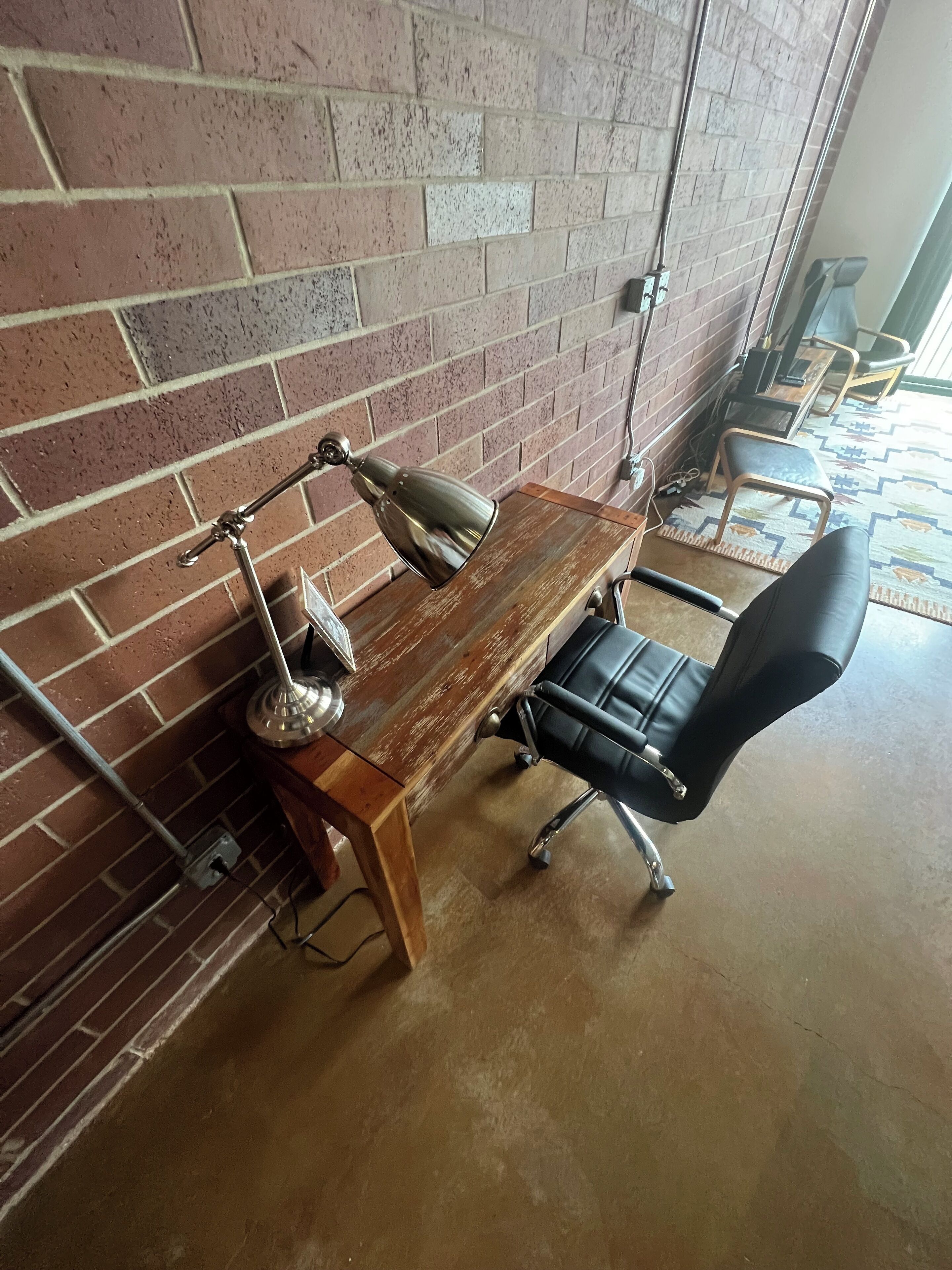 A workspace with a wooden desk, two lamps, a black office chair, and a brick wall.