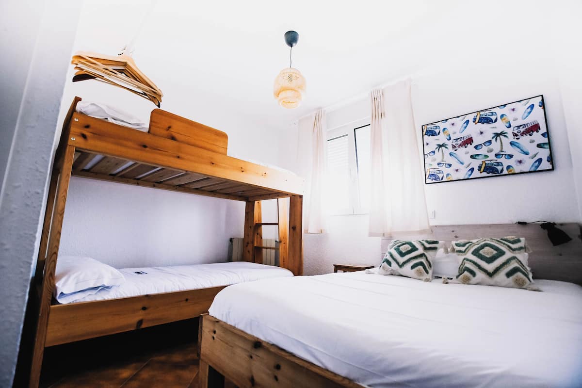 Une chambre avec deux lits superposés, un lit simple, un luminaire suspendu et un mur orné d’une œuvre d’art sur le thème du voyage.