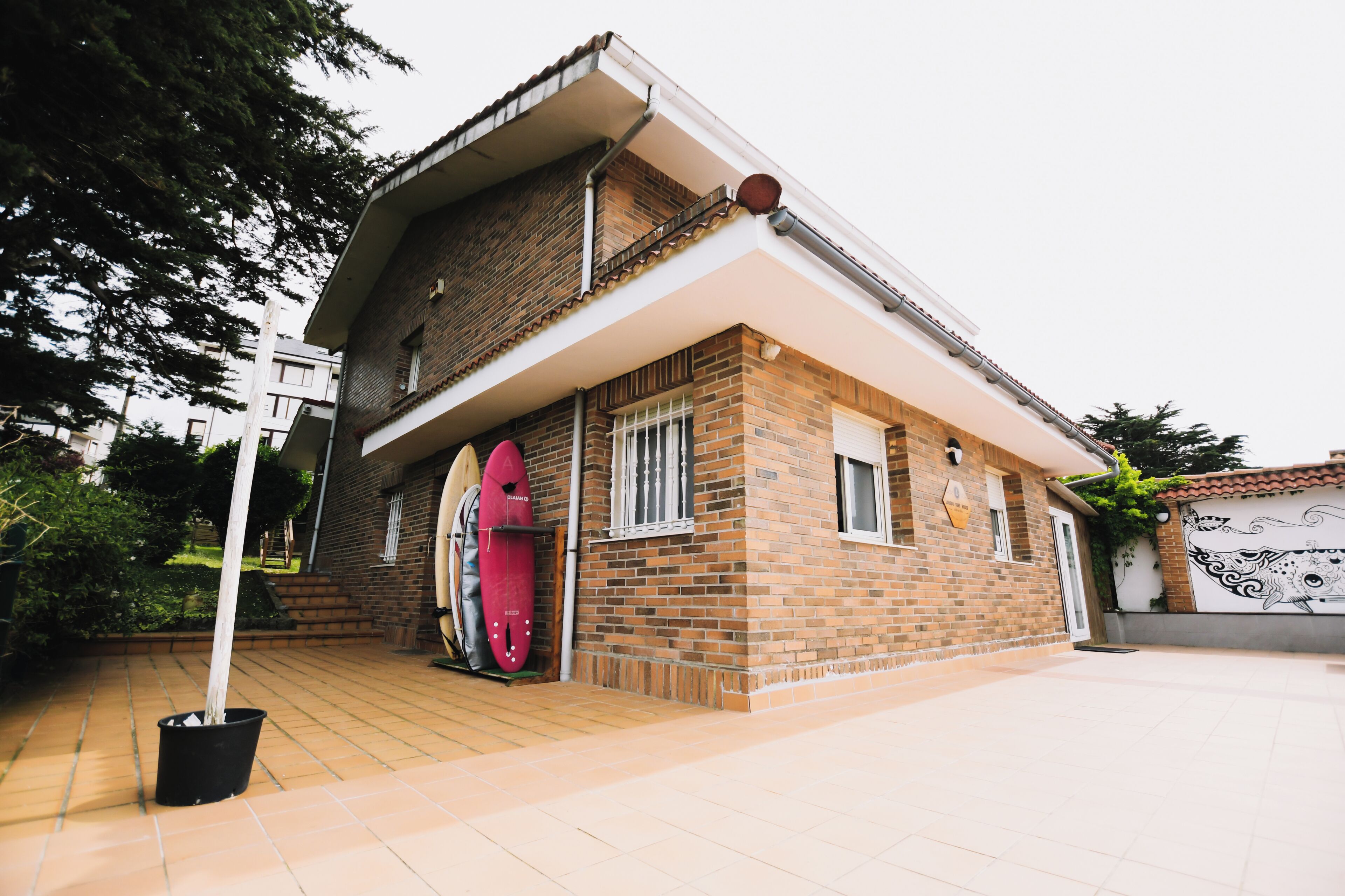 Una casa de dos pisos con fachada de ladrillo, un patio con baldosas y una tabla de surf roja apoyada contra la pared.