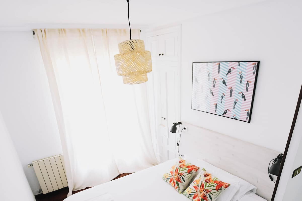 Un lit bien fait, avec du linge de lit blanc et des coussins aux motifs floraux colorés, un luminaire suspendu et un tableau accroché au mur.