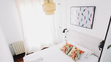 Una cama bien hecha con sábanas blancas y coloridos cojines estampados, una lámpara colgante y un cuadro en la pared.