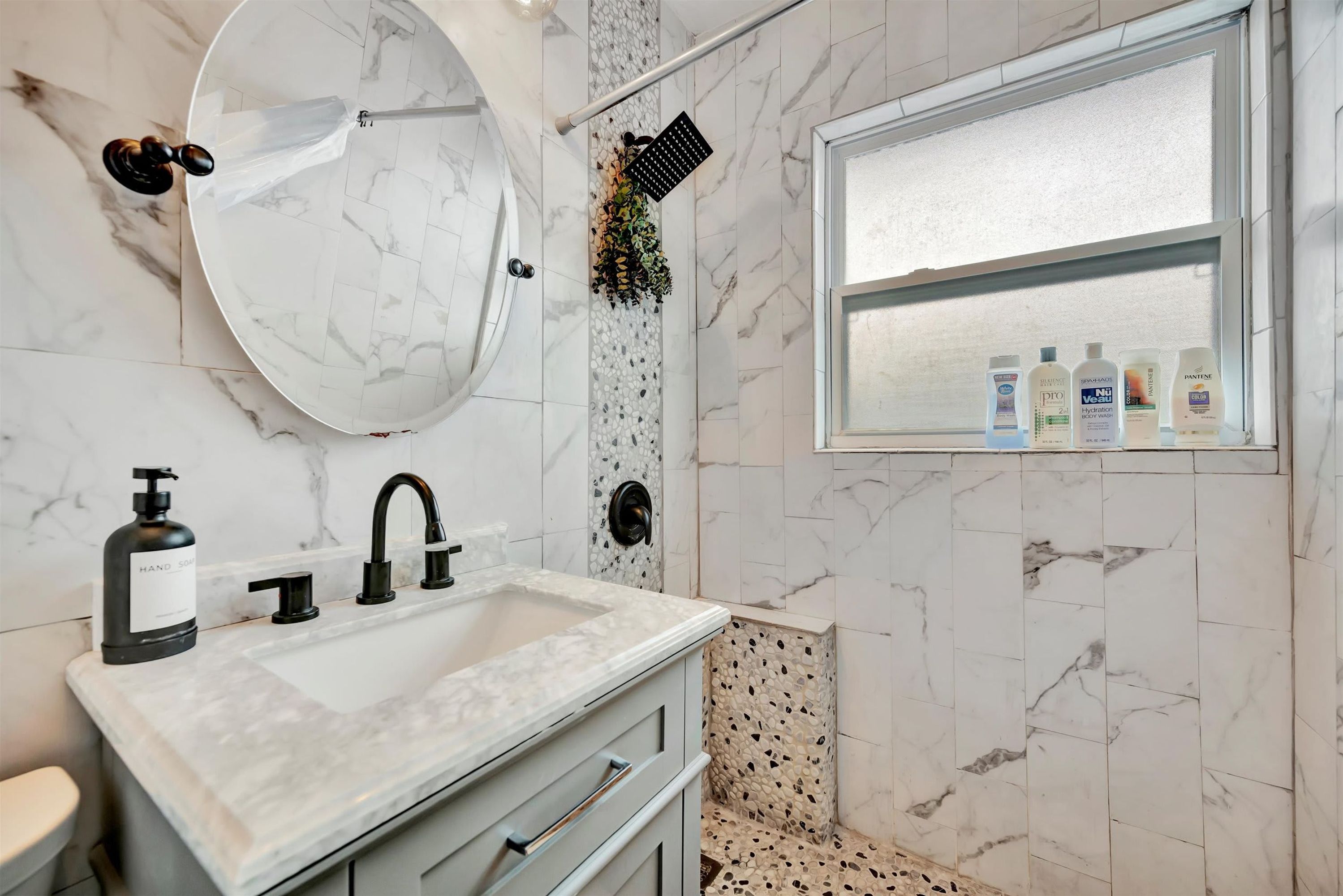 A bathroom with a marble sink, a round mirror, a handheld showerhead, and toiletries on a shelf.