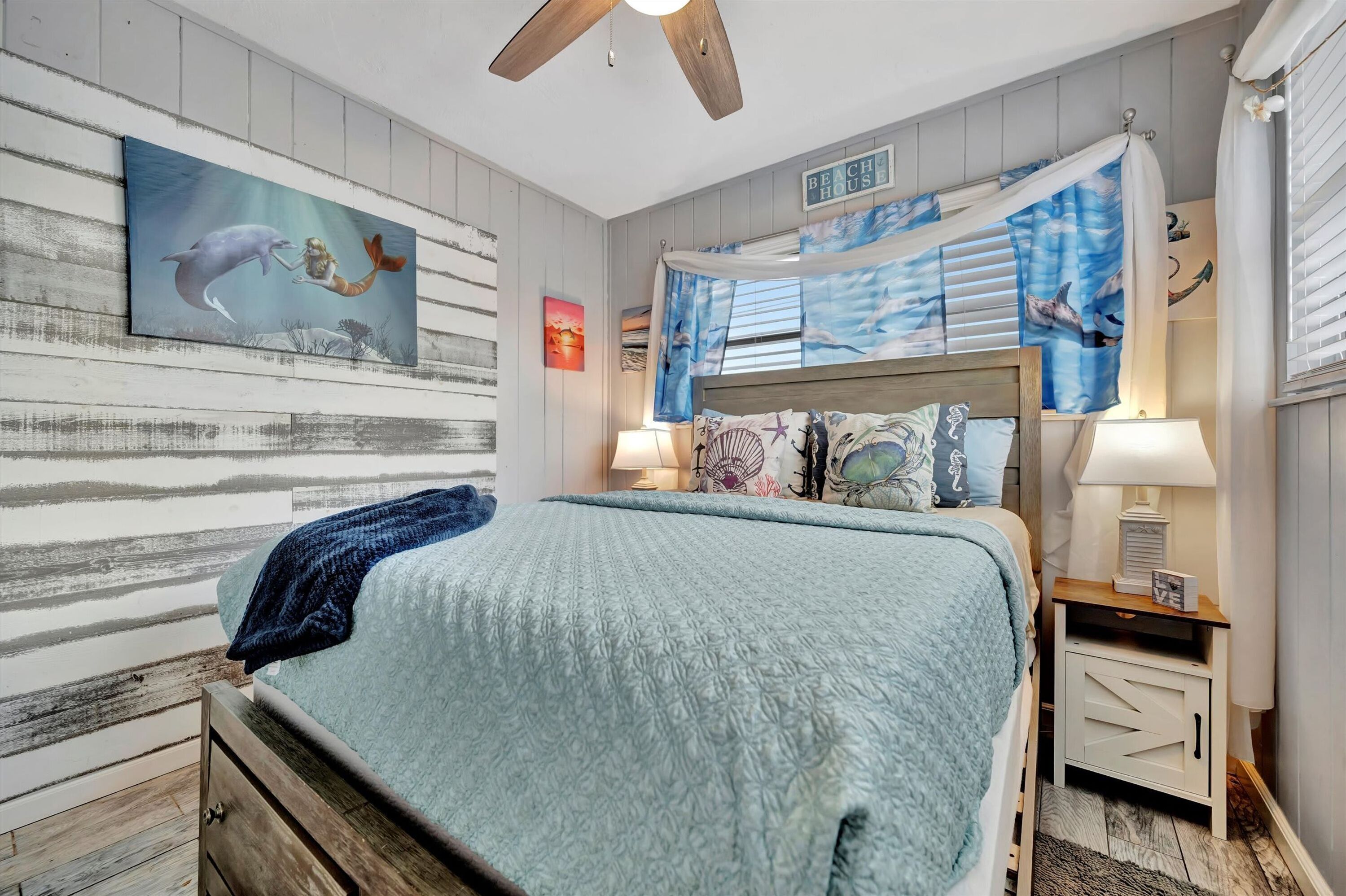 Ein Schlafzimmer mit einem Bett, einem Nachttisch, einem Deckenventilator und einem Wandbild mit Strandmotiv.