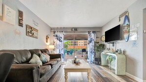 A living room with a brown leather sofa, a wooden coffee table, and a TV on the wall. There are curtains, a surfboard, and a sign that says "Home is where the beach is." - Dolphin Suite 2B Pet Friendly Waterfront Dock (Treasure Island)