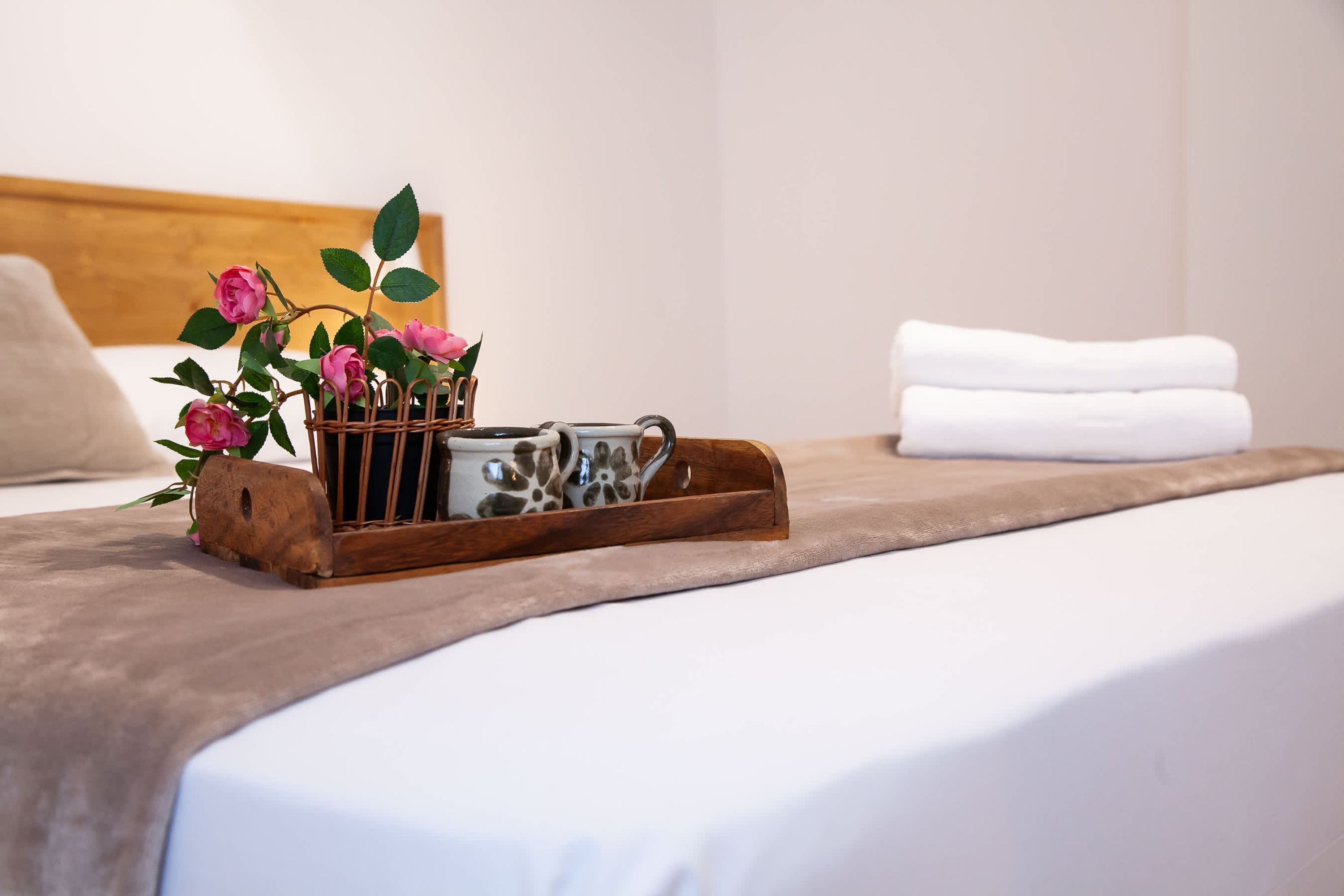 Uma cama bem arrumada com edredom branco, manta marrom e bandeja de madeira com um vaso de flores cor-de-rosa e uma caneca decorativa.