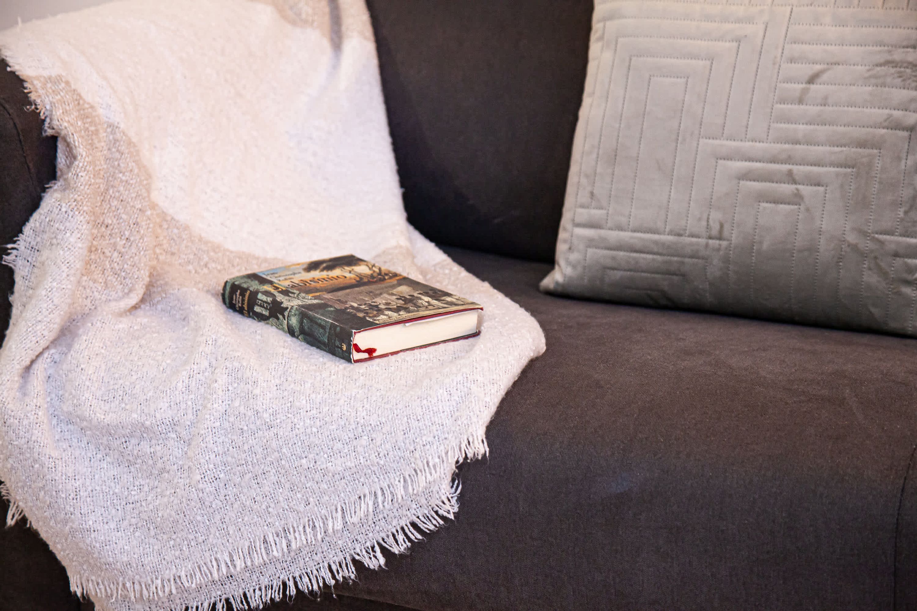 A book on a white knitted blanket on a brown couch with a patterned pillow.