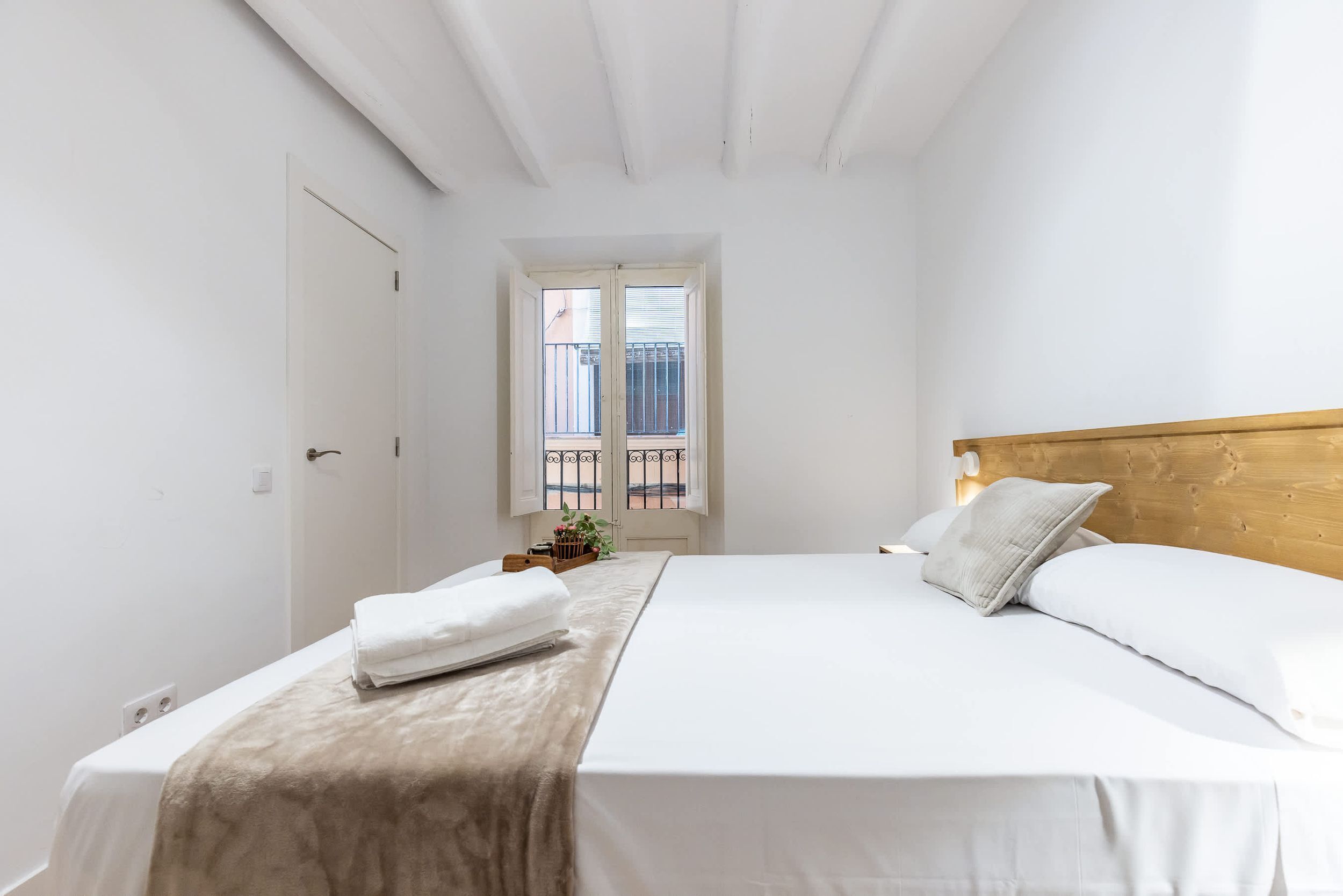 Une chambre à coucher avec un lit, une fenêtre donnant sur un balcon, une tête de lit en bois et une porte blanche.
