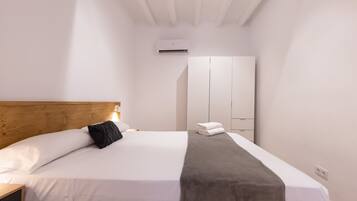 Una cama bien tendida con ropa de cama blanca y gris, un cabecero de madera y una mesita de noche con una lámpara.