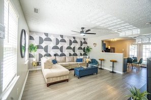 A spacious living room with a large sectional sofa, a blue ottoman, and a kitchen area with bar stools. - 2br each w/ private full bath (Raleigh)