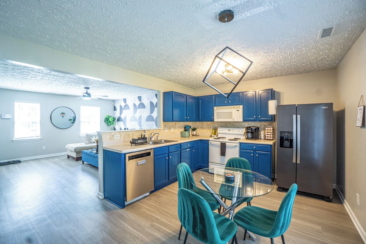 A modern kitchen with blue cabinets, stainless steel appliances, and a glass dining table with teal chairs.