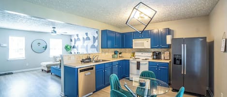 A modern kitchen with blue cabinets, stainless steel appliances, and a glass dining table with teal chairs.