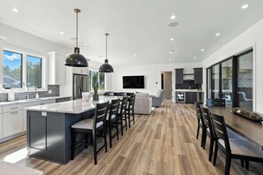 A modern kitchen with a central island, bar stools, a dining area with a TV, and a large sliding glass door to the right. - New Build Baileys Harbor-sunset view & pool table (Baileys Harbor)