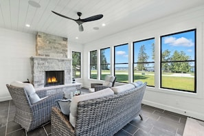 A room with a stone fireplace, wicker furniture, and large windows offering a view of a golf course. - New Build Baileys Harbor-sunset view & pool table (Baileys Harbor)